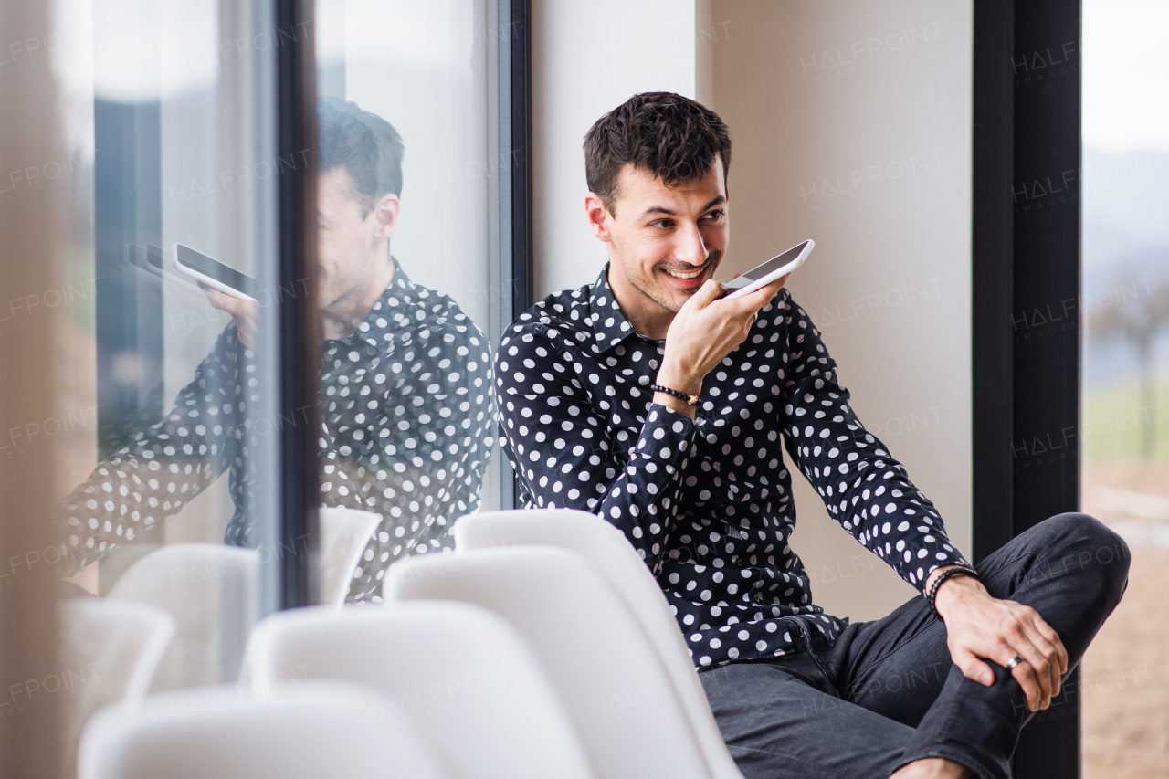 A young man with smartphone sitting by the window, making phone call. Copy space.