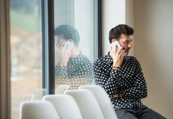 A young man with smartphone sitting by the window, making phone call. Copy space.