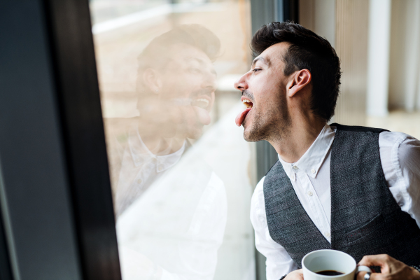 A young man with coffee standing by the window, sticking out tongue.