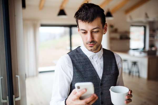 A young man with coffee and smartphone standing indoors at home.