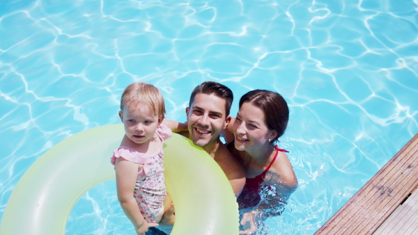 Happy young family with small daughter in swimming pool outdoors in backyard garden, looking at camera.