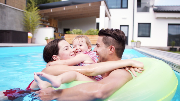 Happy young family with small daughter in swimming pool outdoors in backyard garden, looking at camera.