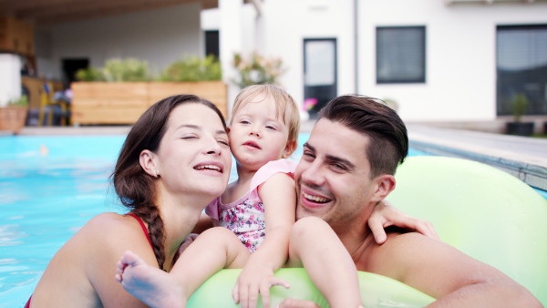 Happy young family with small daughter in swimming pool outdoors in backyard garden, looking at camera.