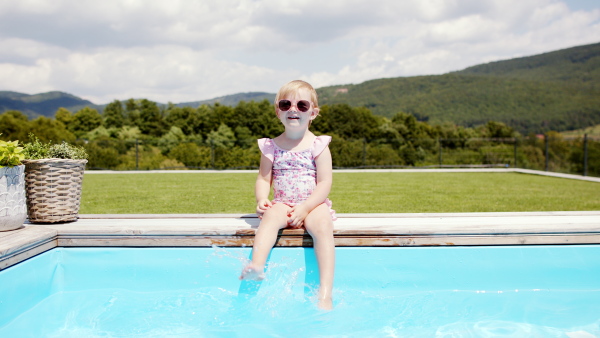 Portrait of small toddler girl sitting by swimming pool outdoors in backyard garden, looking at camera.