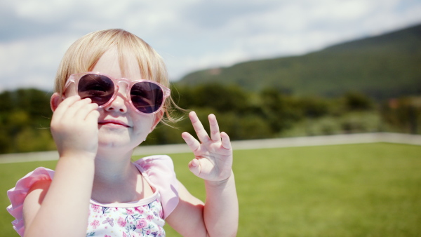 Portrait of small toddler girl sitting in backyard garden, playing with sunglasses.