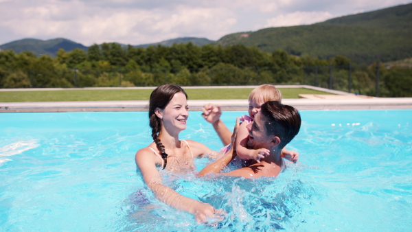 Happy young family with small daughter in swimming pool outdoors in backyard garden, looking at camera.