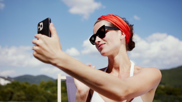 Young woman sitting by swimming pool outdoors in backyard garden, taking selfie with smartphone.