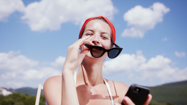 A young woman sitting by swimming pool with smartphone outdoors in backyard garden, looking at camera.