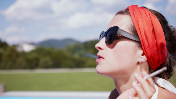 Young woman with drink sitting by swimming pool outdoors in backyard garden, smoking and relaxing.