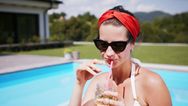 Young woman with drink sitting by swimming pool outdoors in backyard garden, relaxing and looking at camera.