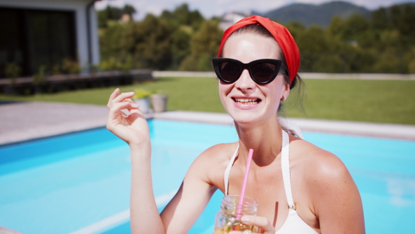 Young woman with drink sitting by swimming pool outdoors in backyard garden, relaxing and looking at camera.