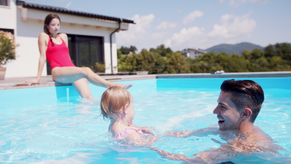 Happy young family with small daughter in swimming pool outdoors in backyard garden.