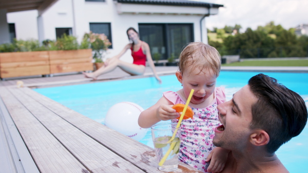 Portrait of small girl with father drinking lemonade in swimming pool outdoors in backyard garden.