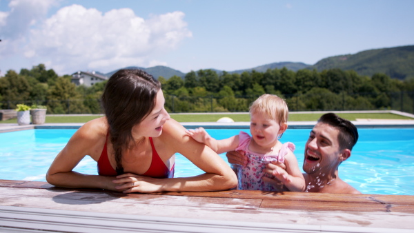 Happy young family with small daughter in swimming pool outdoors in backyard garden.