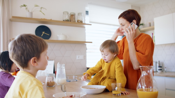 Busy mother with small children in kitchen in the morning at home, using smartphone when eating breakfast.