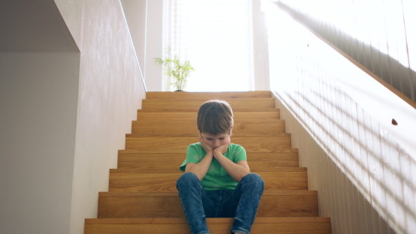 Front view portrait of sad small boy sitting on stairs at home, head in hands