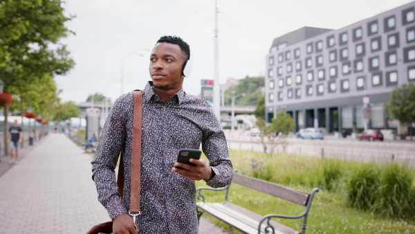 Young black man commuter outdoors in city, walking with headphones and smartphone.