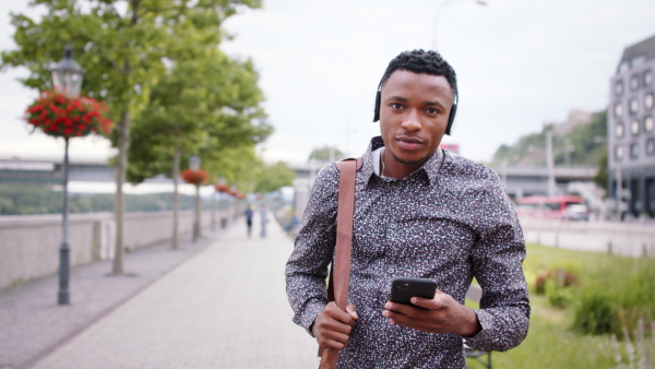 Young black man commuter outdoors in city, walking with headphones and smartphone.