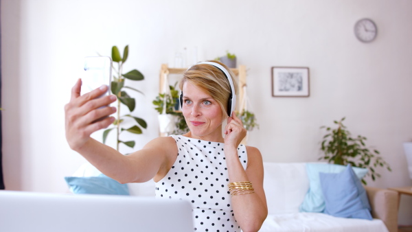 Cheerful young businesswoman with headphones and telephone indoors in home office, taking selfie.