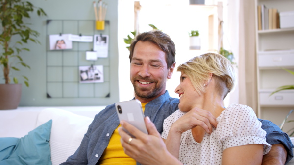 Laughing couple in love sitting on sofa indoors at home, using smartphone.