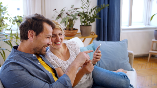 Happy young couple with smartphone sitting indoors at home, laughing.