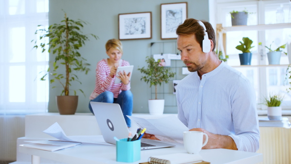 Mature architect with headphones, laptop and partner in the background indoors in home office, working.