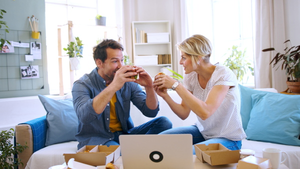 Front view of happy couple sitting on sofa indoors at home, eating hamburgers.