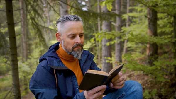 Happy mature man reading book outdoors in forest in nature, sitting.