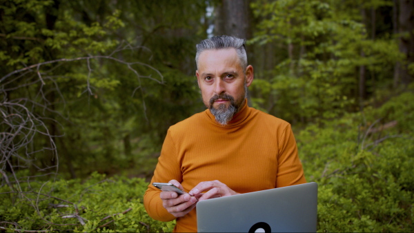 Front view of mature man with laptop and smartphone in forest in nature, outdoor office concept.