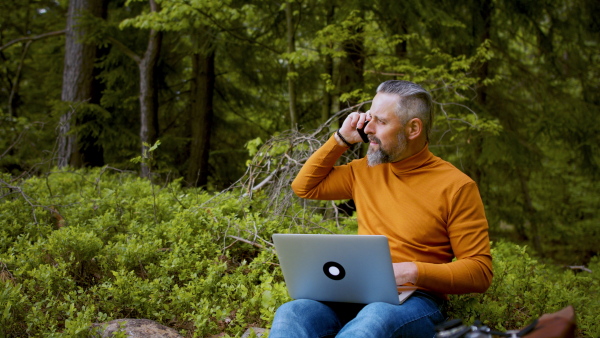 Front view of mature man with laptop and smartphone working in forest in nature, outdoor office concept.