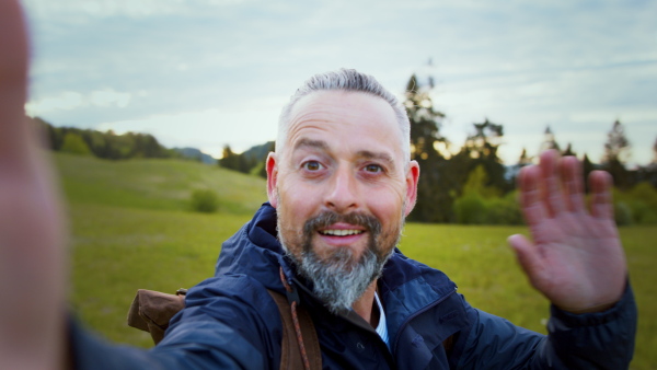 Mature man with backpack outdoors on grassland in nature, taking selfie with smartphone.