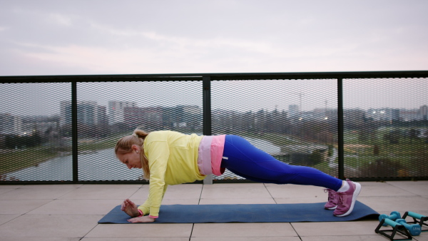 Side view of young woman in yellow tracksuit exercising outside on terrace.