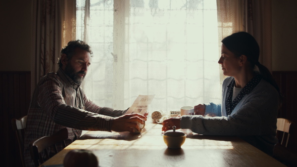 Side view of unhappy poor mature couple with coffee reading newspapers indoors at home, poverty concept.