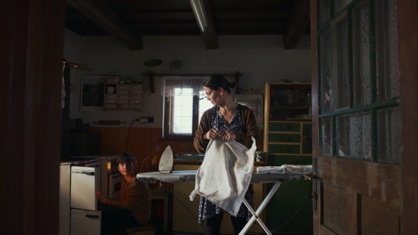 Front view of poor mother with small daughter indoors at home, ironing. Poverty concept.