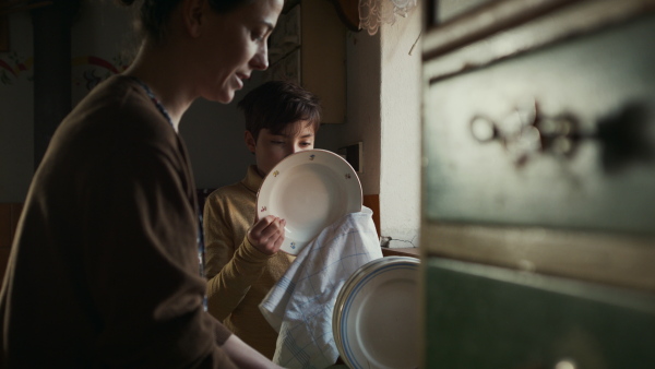 Portrait of poor mature mother and small daughter washing dishes indoors at home, poverty concept.