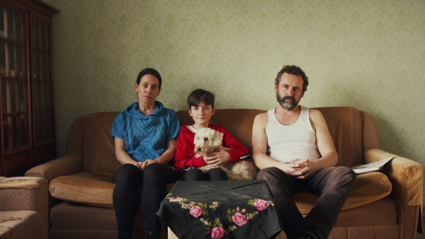 Portrait of poor mature couple with small daughter indoors on sofa at home, looking at camera. Poverty concept.