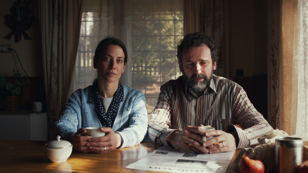 Front view of unhappy poor mature couple with coffee reading newspapers indoors at home, looking at camera. Poverty concept.