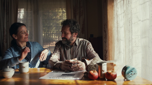 Front view of humble poor mature couple with coffee indoors at home, looking at camera. Poverty concept.