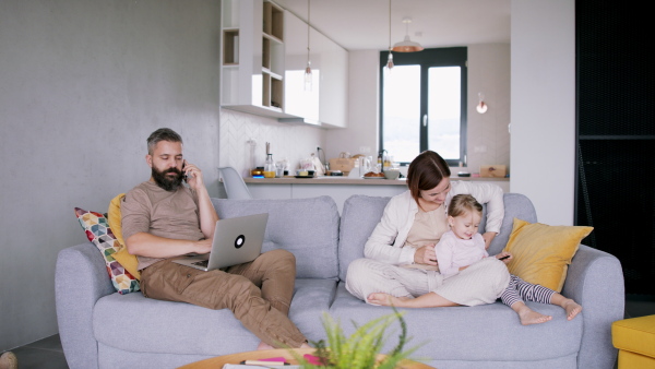 Father sitting on sofa and working from home next to his wife and daughter. Homeoffice concept.