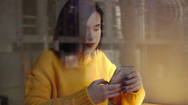 Happy young woman sitting in cafe, using smartphone. Shot through glass.