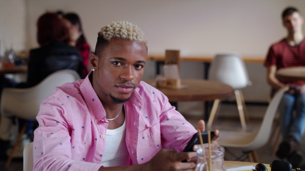 Front view of young african american man with smartphone sitting in cafe, looking at camera. Slow motion.