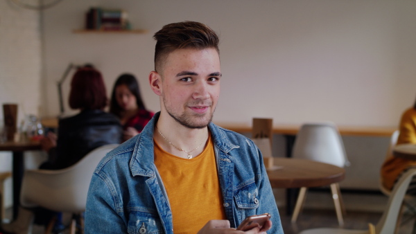 Front view of young man with smartphone sitting in cafe, looking at camera.