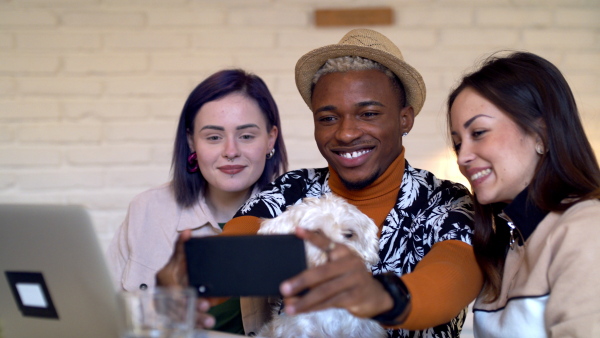 Group of cheerful young friends with dog and smartphone sitting in cafe, taking selfie.