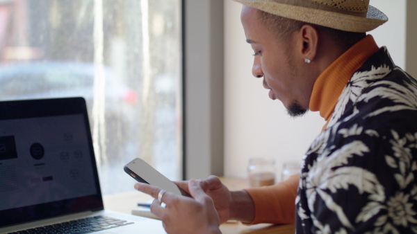 Cheerful young black man sitting and working in cafe, using laptop and smartphone.