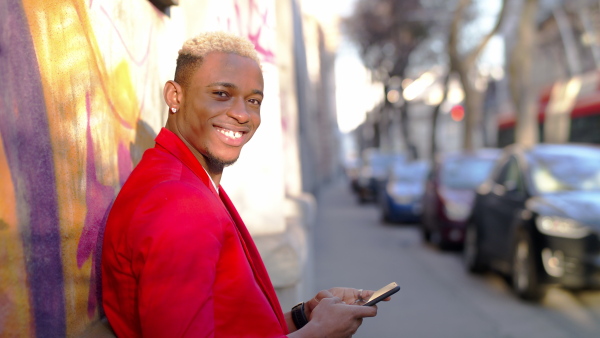 Side view portrait of happy young black man standing outdoors on street, using smartphone.