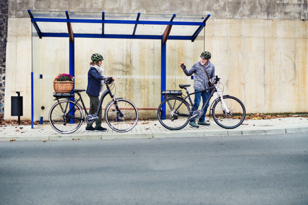 Active senior couple with helmets and electrobikes standing outdoors on a road in town.