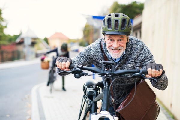 Active senior couple with helmets and electrobikes standing outdoors on a road in town.