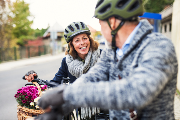 Active senior couple with helmets and electrobikes standing outdoors on a road in town.