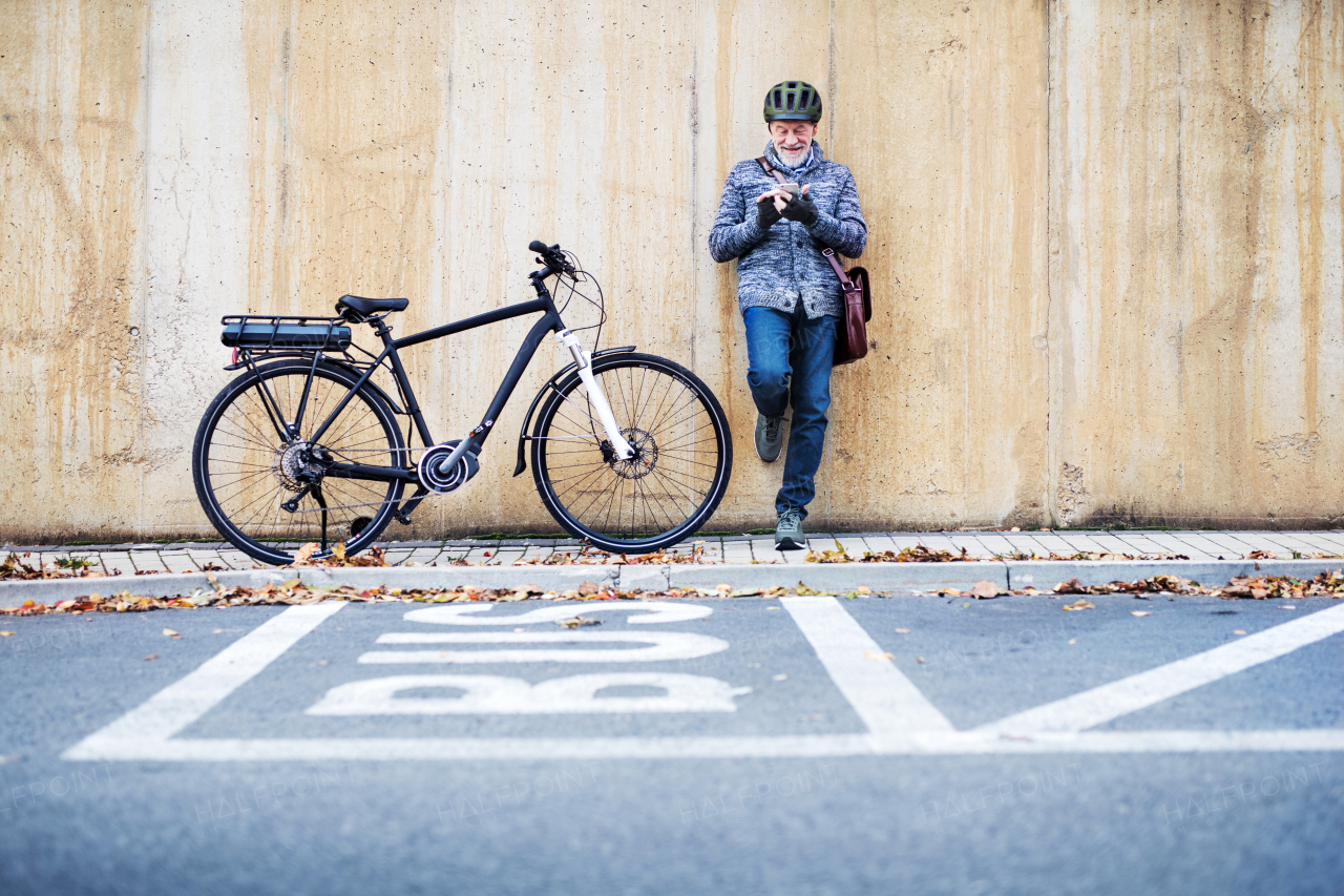 Active senior man with electrobike standing outdoors in town, leaning against a concrete wall and using smartphone. Copy space.