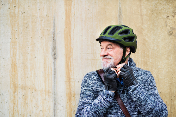 Active senior man standing outdoors against a concrete wall in town, putting on bicycle helmet. Copy space.